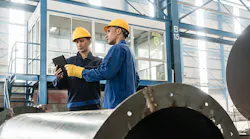 A pair of worker stand over a tablet in a factory.