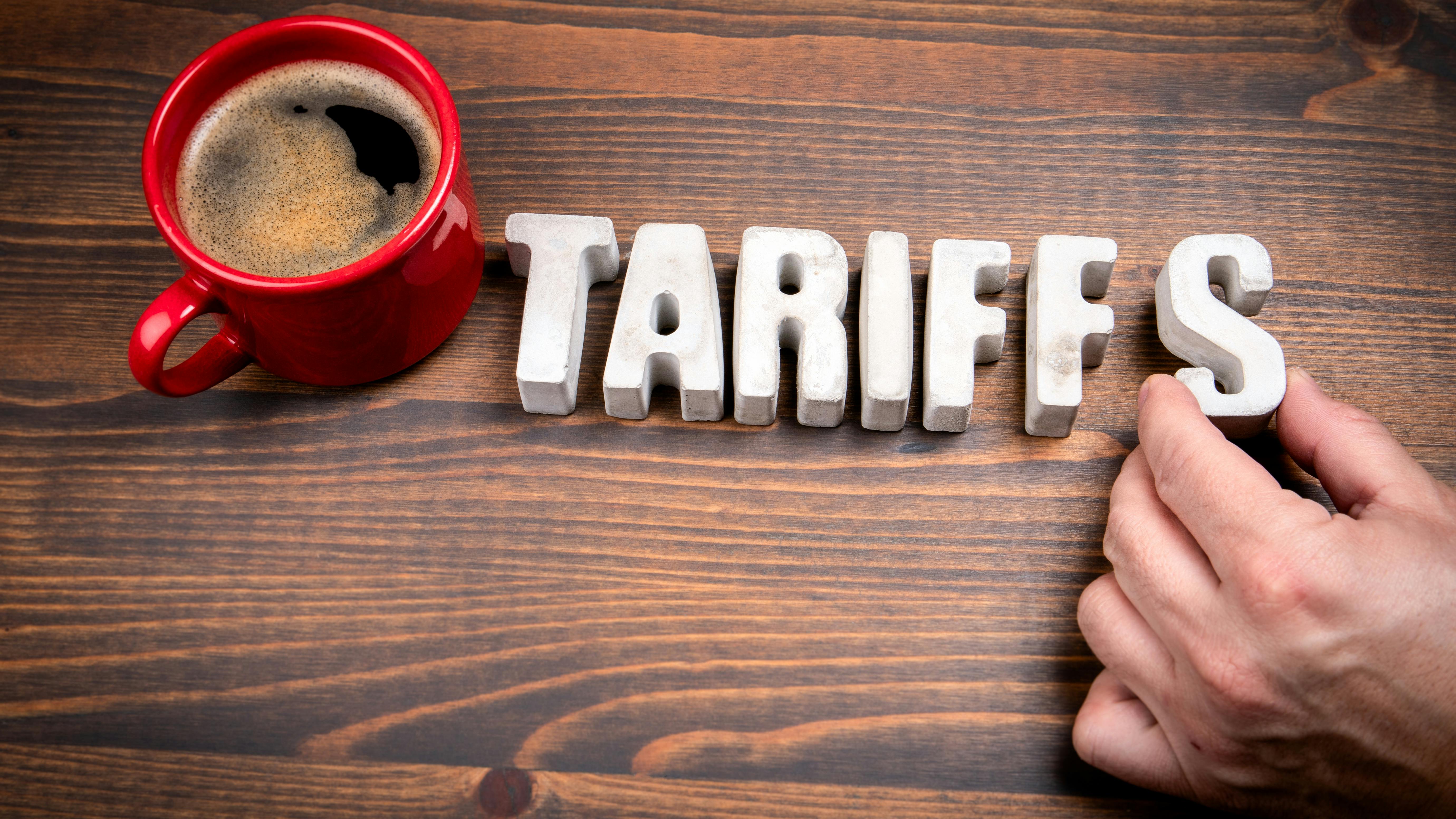 Concrete alphabet letters spelling 'Tariffs' and coffee mug on dark wooden texture background.