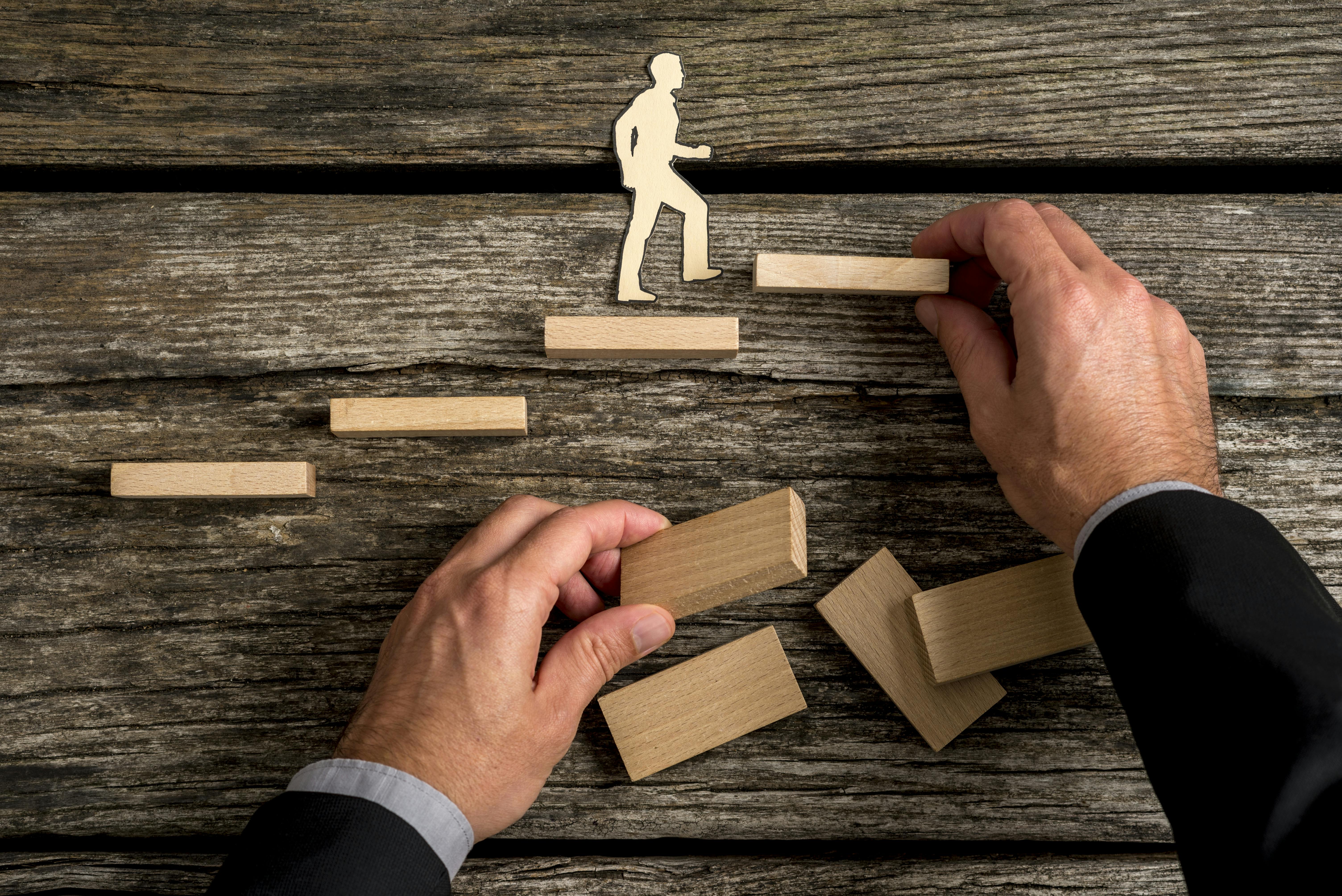 Silhouette cutouts of a man walking up steps. The hand of a businessman is building the stairs for him.